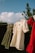 A line of children's clothes hanging on a clothesline outdoors. The dresses are in olive green, cream, and red colors, gently swaying in the breeze. The background features a clear blue sky with fluffy clouds and some green foliage.