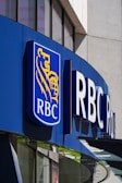 A modern building facade features a prominent logo that includes a blue background with a golden lion and globe. The letters 'RBC' are boldly displayed next to the logo, indicating a business establishment. The structure is made of glass and metal, with reflections visible on the windows.