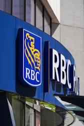 A modern building facade features a prominent logo that includes a blue background with a golden lion and globe. The letters 'RBC' are boldly displayed next to the logo, indicating a business establishment. The structure is made of glass and metal, with reflections visible on the windows.
