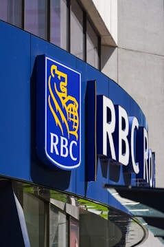 A modern building facade features a prominent logo that includes a blue background with a golden lion and globe. The letters 'RBC' are boldly displayed next to the logo, indicating a business establishment. The structure is made of glass and metal, with reflections visible on the windows.