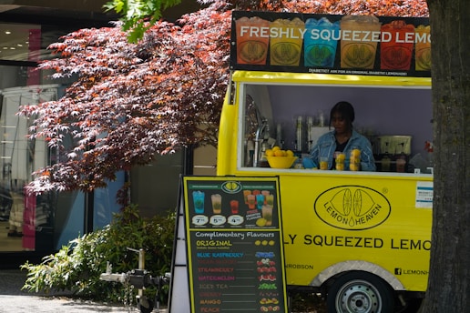 A vibrant yellow lemonade stand with signage advertising freshly squeezed lemonade. The stand displays various drink options and is adorned with a basket of lemons. A person is preparing a drink inside the stand. A colorful menu board with different flavors is positioned outside. The scene is set against the backdrop of a leafy, red-hued tree and a modern building.