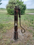 An old, weathered hand-operated water pump stands in a grassy open area. The pump has a rusty red and brownish exterior, and is situated in front of dense green bushes. The ground around the pump is dry and sparse, with patches of grass and dirt visible.