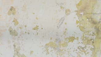 Large patches of peeling paint expose various shades of green and yellow on a weathered wall surface. The texture is rough with some cracks and flaking, creating an abstract pattern.