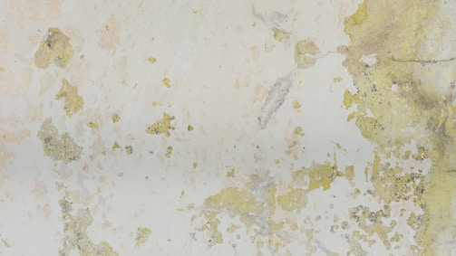 Large patches of peeling paint expose various shades of green and yellow on a weathered wall surface. The texture is rough with some cracks and flaking, creating an abstract pattern.