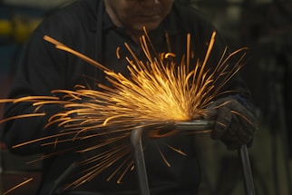 Sparks fly as a worker uses a grinder on a metal piece, creating a burst of bright orange light against a dark background. The worker wears protective gloves and clothing while focusing intently on the task.