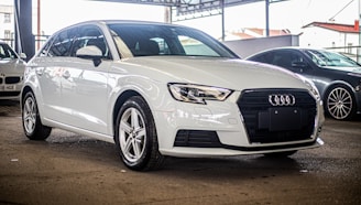 A sleek, white Audi car is parked in a covered area, with other vehicles visible in the background. The car is positioned at an angle, showcasing its front and side profile. The environment suggests a dealership or showroom setting with ample lighting.