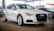 A sleek, white Audi car is parked in a covered area, with other vehicles visible in the background. The car is positioned at an angle, showcasing its front and side profile. The environment suggests a dealership or showroom setting with ample lighting.