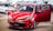 A bright red car with sporty design details is parked indoors with its front doors open. The car has a sleek exterior, highlighted by aerodynamic features and modern headlights. Several other cars are parked in the background, suggesting a showroom or garage setting.
