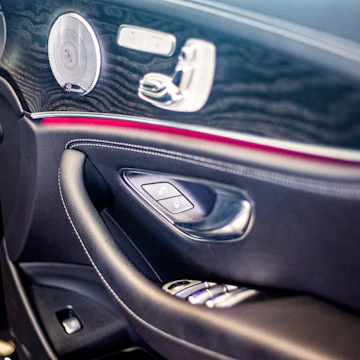 The image features the interior door panel of a luxury car, showcasing a sleek and modern design. It includes an elegant combination of materials such as leather and wood grain finish. Visible elements include a high-quality speaker with chrome accents, seat adjustment buttons, and a window control panel.