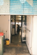 A view through an open roller shutter door leading into a room with glass doors. Inside, there is a person working near metal racks filled with trays. The area outside and immediately inside the entrance is clean, with a mop, bucket, trash bin, and multiple mats on the floor.