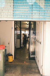 A view through an open roller shutter door leading into a room with glass doors. Inside, there is a person working near metal racks filled with trays. The area outside and immediately inside the entrance is clean, with a mop, bucket, trash bin, and multiple mats on the floor.