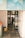 A view through an open roller shutter door leading into a room with glass doors. Inside, there is a person working near metal racks filled with trays. The area outside and immediately inside the entrance is clean, with a mop, bucket, trash bin, and multiple mats on the floor.