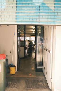 A view through an open roller shutter door leading into a room with glass doors. Inside, there is a person working near metal racks filled with trays. The area outside and immediately inside the entrance is clean, with a mop, bucket, trash bin, and multiple mats on the floor.