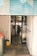 A view through an open roller shutter door leading into a room with glass doors. Inside, there is a person working near metal racks filled with trays. The area outside and immediately inside the entrance is clean, with a mop, bucket, trash bin, and multiple mats on the floor.