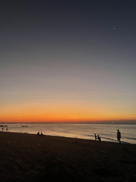 The image captures a serene beach scene at sunset, with an expanse of sand leading to calm waters. The sky transitions from deep blue at the top to a warm orange and yellow near the horizon. Silhouettes of people are visible on the shoreline, some walking and others sitting, creating a peaceful and reflective atmosphere.