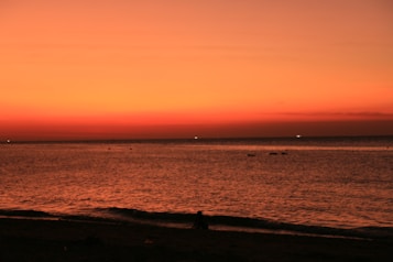 The image features a serene beach scene during sunset, with a vast expanse of water meeting the horizon. The sky is painted in warm hues of orange and red, creating a dramatic and calming effect. A silhouette of a person can be seen sitting on the shore, adding a human element to the natural beauty.
