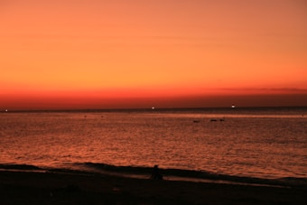 The image features a serene beach scene during sunset, with a vast expanse of water meeting the horizon. The sky is painted in warm hues of orange and red, creating a dramatic and calming effect. A silhouette of a person can be seen sitting on the shore, adding a human element to the natural beauty.