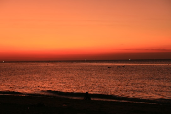The image features a serene beach scene during sunset, with a vast expanse of water meeting the horizon. The sky is painted in warm hues of orange and red, creating a dramatic and calming effect. A silhouette of a person can be seen sitting on the shore, adding a human element to the natural beauty.