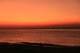 The image features a serene beach scene during sunset, with a vast expanse of water meeting the horizon. The sky is painted in warm hues of orange and red, creating a dramatic and calming effect. A silhouette of a person can be seen sitting on the shore, adding a human element to the natural beauty.