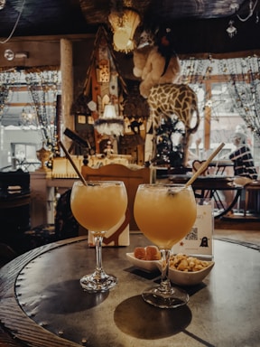A cozy, eclectic interior with warm lighting features two tropical cocktails with straws on a round table. Next to the drinks are small bowls of snacks. The background includes decorative elements like plush giraffes, hanging lamps, and beaded curtains, contributing to a whimsical and inviting atmosphere.
