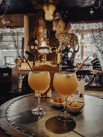 A cozy, eclectic interior with warm lighting features two tropical cocktails with straws on a round table. Next to the drinks are small bowls of snacks. The background includes decorative elements like plush giraffes, hanging lamps, and beaded curtains, contributing to a whimsical and inviting atmosphere.