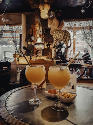 A cozy, eclectic interior with warm lighting features two tropical cocktails with straws on a round table. Next to the drinks are small bowls of snacks. The background includes decorative elements like plush giraffes, hanging lamps, and beaded curtains, contributing to a whimsical and inviting atmosphere.
