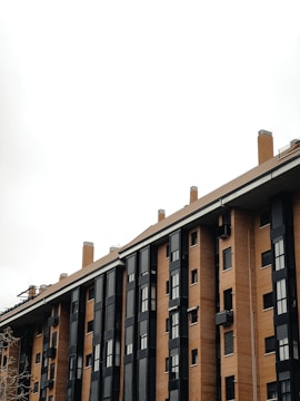 A multi-story residential building with a modern design featuring large, black-framed windows and orange-brown brickwork. The structure includes multiple chimneys and a flat roof, set against an overcast sky.