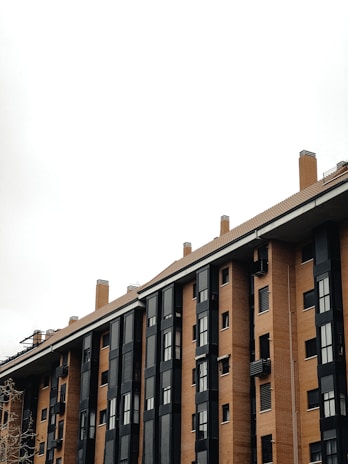 A multi-story residential building with a modern design featuring large, black-framed windows and orange-brown brickwork. The structure includes multiple chimneys and a flat roof, set against an overcast sky.