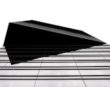 A modern architectural structure with a sharp, geometric design. The building features a series of parallel lines that create a sense of depth and complexity. The high contrast between the black elements and the white background emphasizes the clean, minimalist style.