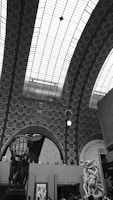 An indoor architectural scene with an intricate arched ceiling composed of a patterned grid design. There are large sculptural artworks, including a prominent globe held by human figures and a classical statue. The setting appears to be a museum or gallery space, with visitors observing the art.