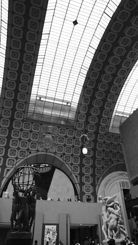 An indoor architectural scene with an intricate arched ceiling composed of a patterned grid design. There are large sculptural artworks, including a prominent globe held by human figures and a classical statue. The setting appears to be a museum or gallery space, with visitors observing the art.