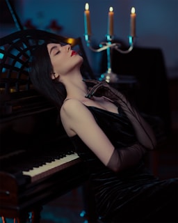 A woman in an elegant black dress leans back against a piano with a relaxed and contemplative expression. Her eyes are closed, and she gently touches her neck with one hand wearing a sheer glove. In the background, a candelabra with lit candles casts a warm glow, creating a moody and intimate atmosphere.
