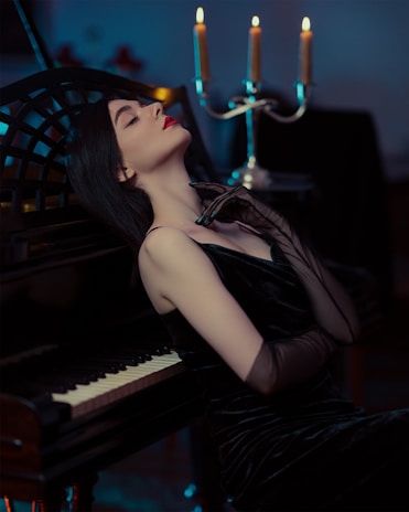 A woman in an elegant black dress leans back against a piano with a relaxed and contemplative expression. Her eyes are closed, and she gently touches her neck with one hand wearing a sheer glove. In the background, a candelabra with lit candles casts a warm glow, creating a moody and intimate atmosphere.