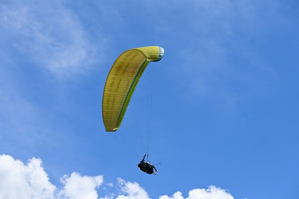 A paraglider with a yellow canopy is flying against a clear blue sky. The paraglider is positioned slightly above some white clouds, creating a sense of freedom and adventure.
