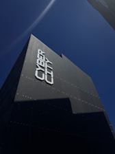 A tall, modern building is captured from a low angle against a clear blue sky. The facade of the building reads 'CYBER CITY' in large, bold letters. The building's surface appears sleek and dark, with minimal reflective features.