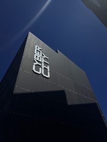 A tall, modern building is captured from a low angle against a clear blue sky. The facade of the building reads 'CYBER CITY' in large, bold letters. The building's surface appears sleek and dark, with minimal reflective features.