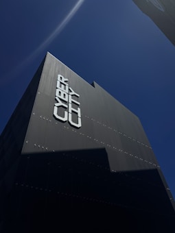 A tall, modern building is captured from a low angle against a clear blue sky. The facade of the building reads 'CYBER CITY' in large, bold letters. The building's surface appears sleek and dark, with minimal reflective features.