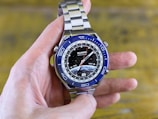 A hand is holding a modern wristwatch with a blue and silver bezel and a metal strap. The watch face displays multiple time zones with city abbreviations on its outer rim and a detailed image of the Earth at the center. The brand 'Huawei Watch Ultimate' is visible on the dial.