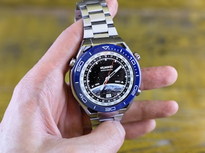A hand is holding a modern wristwatch with a blue and silver bezel and a metal strap. The watch face displays multiple time zones with city abbreviations on its outer rim and a detailed image of the Earth at the center. The brand 'Huawei Watch Ultimate' is visible on the dial.