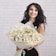 A woman with long, wavy black hair is smiling while holding a large bouquet of daisies. She is wearing a black and beige sleeveless dress and standing against a light-colored brick wall.