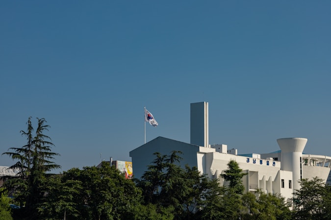 A modern white building with a prominent South Korean flag on a pole. The structure is surrounded by lush green trees. The sky is clear and blue, and there is Korean text visible on the building.