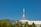 A tall orange and white communication tower rises above a mix of modern buildings and lush green trees. The clear blue sky provides a crisp backdrop, enhancing the urban and natural elements. A gently sloping hill is visible in the distance.
