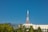 A tall orange and white communication tower rises above a mix of modern buildings and lush green trees. The clear blue sky provides a crisp backdrop, enhancing the urban and natural elements. A gently sloping hill is visible in the distance.