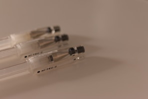 A close-up of three transparent pens with metallic tips and black caps laying side by side on a smooth, light-colored surface. The pens are positioned diagonally and the lighting creates a soft shadow.