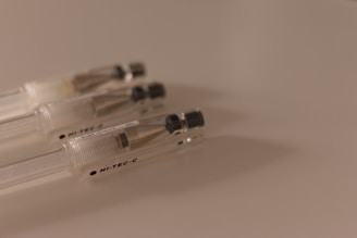 A close-up of three transparent pens with metallic tips and black caps laying side by side on a smooth, light-colored surface. The pens are positioned diagonally and the lighting creates a soft shadow.