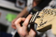 A close-up view of a person's hand playing a guitar, focusing on the neck and tuning pegs. The image emphasizes the strumming fingers and the details of the guitar headstock.
