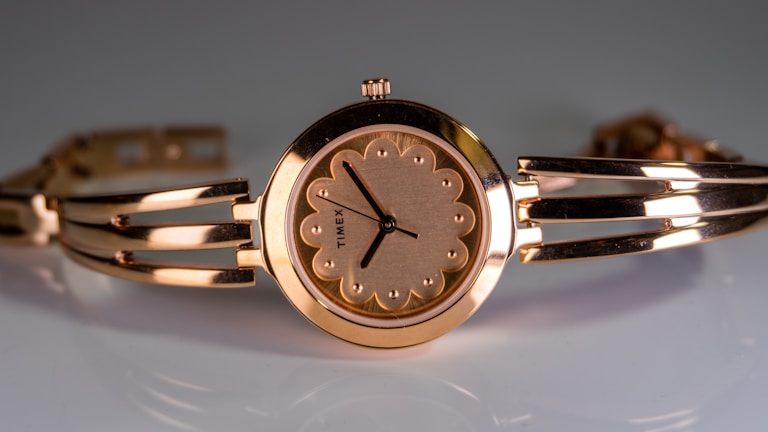 An elegant wristwatch with a gold-toned bracelet and a circular face showcasing a floral-inspired design on the dial. The hands are thin and simple, with a brand name subtly engraved on the face. The metallic sheen of the watch adds a luxurious and sophisticated touch.