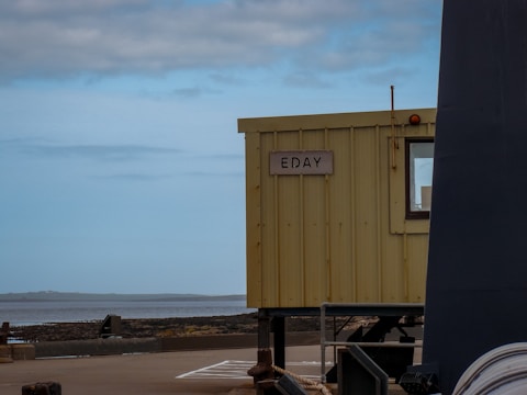 A small, yellow building with a sign reading 'EDAY' is positioned near a body of water. The sky is overcast, and the area around the building appears to be a pier or dockside, with visible concrete and metal structures.