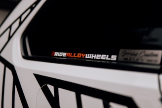 A close-up of a car accent showcasing a decal or sticker that reads 'RIDEALLOYWHEELS' in a bold font. Part of the vehicle is visible with contrasting black and white geometric patterns on the body.