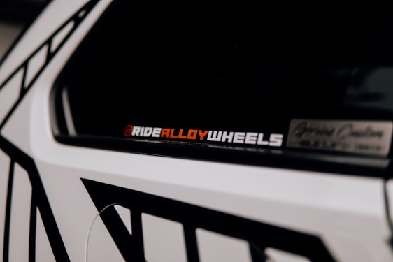 A close-up of a car accent showcasing a decal or sticker that reads 'RIDEALLOYWHEELS' in a bold font. Part of the vehicle is visible with contrasting black and white geometric patterns on the body.